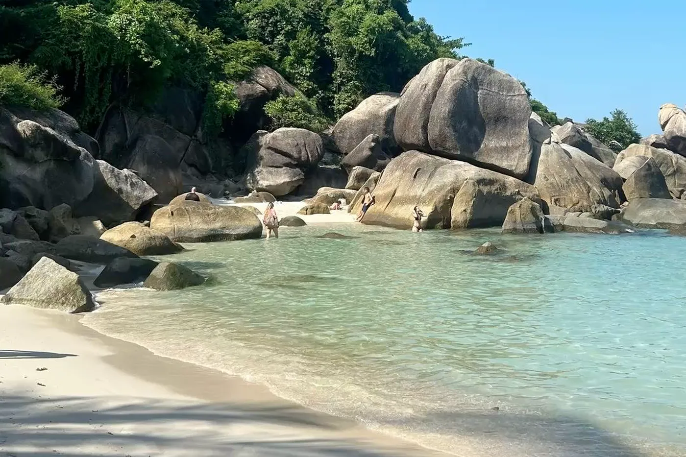 People wading among large granite boulders in a clear turquoise cove, jungle on the rocks above and white sand in the foreground.
