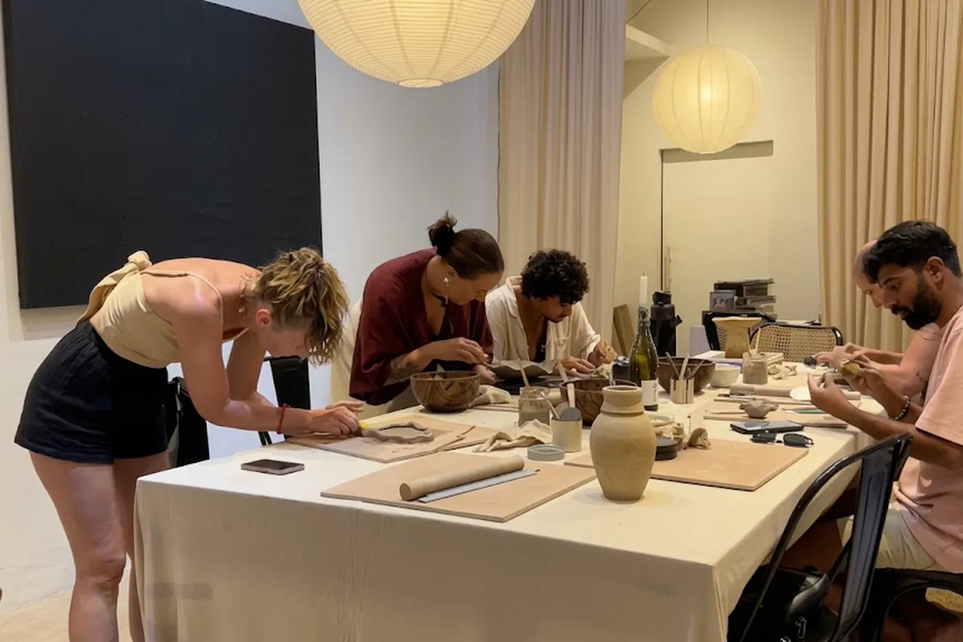 Four people shaping clay together at a long table lit by paper pendant lamps, pots and tools laid out in front of them.