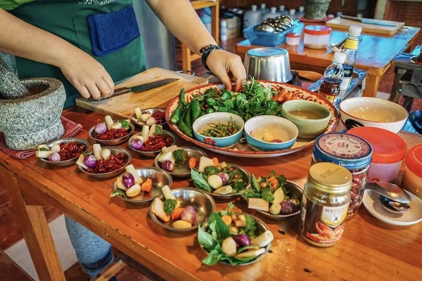 Hands chopping vegetables at a wooden table covered with bowls of prepared Thai ingredients, fresh herbs and spice pastes.
