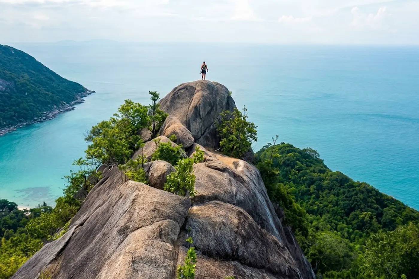 A hiker standing on a granite peak above the jungle, a turquoise cove on one side and open sea on the other.