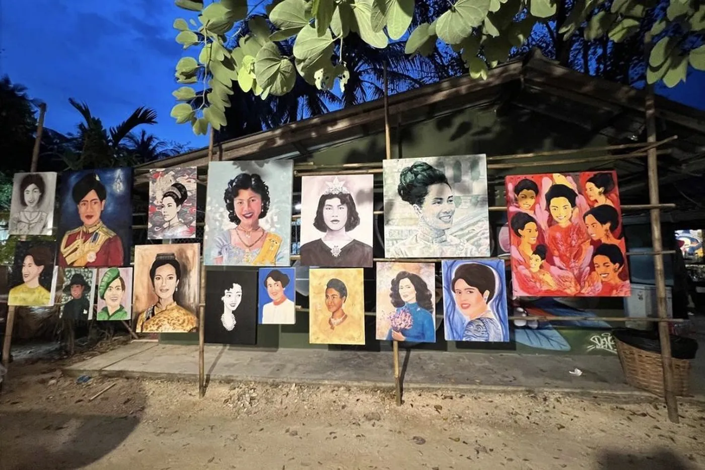 Painted portraits hung across the front of a market stall in the evening, foliage overhead against a deep blue sky.