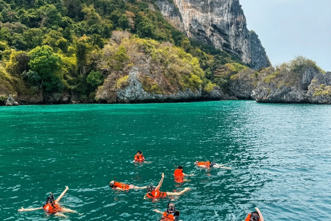 Turquoise water below a steep limestone cliff covered in jungle, with several snorkellers in orange flotation vests near the rock.