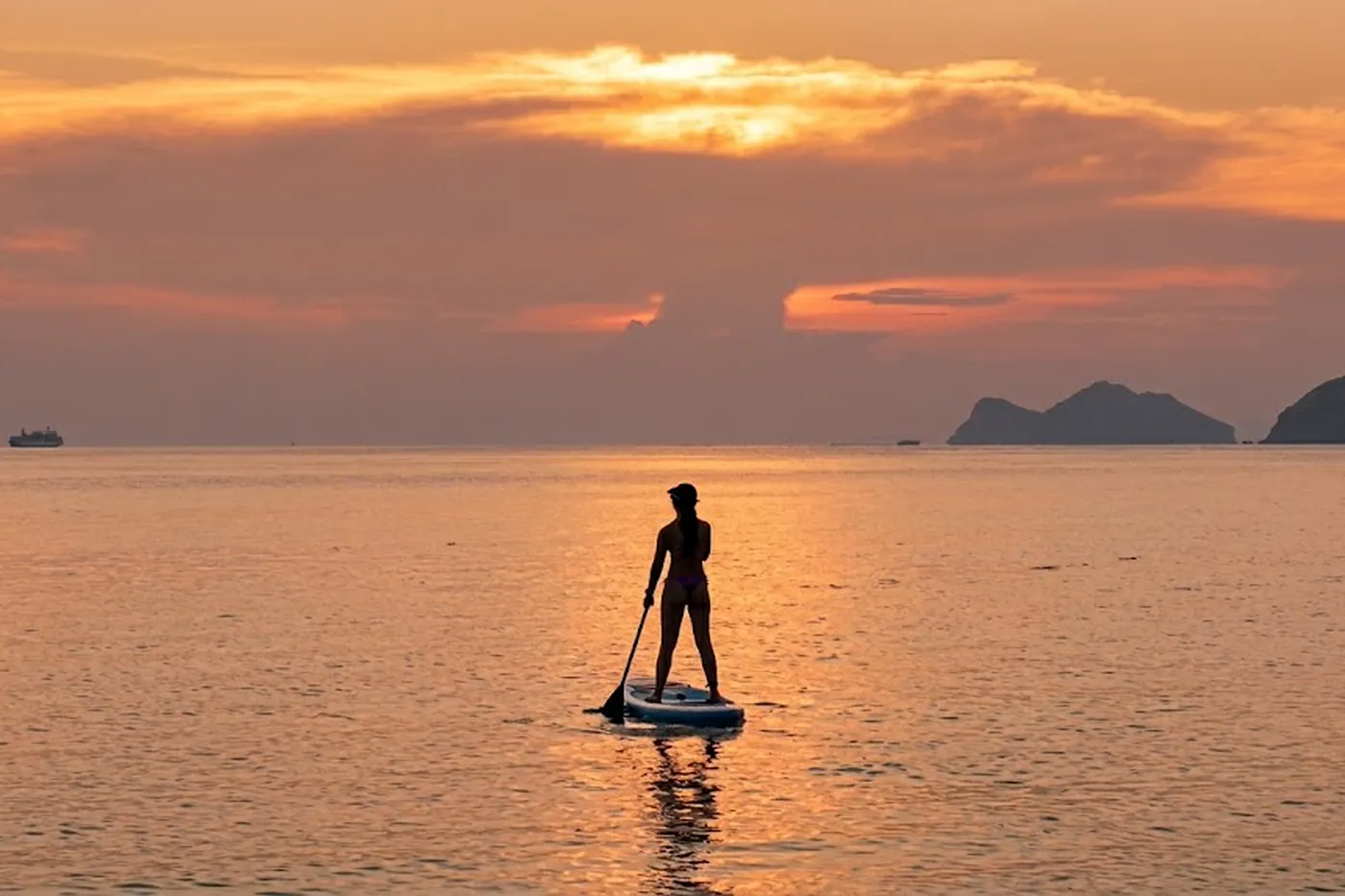 A paddleboarder in silhouette on flat water at sunset, mountains and a passing boat on the horizon under an orange sky.