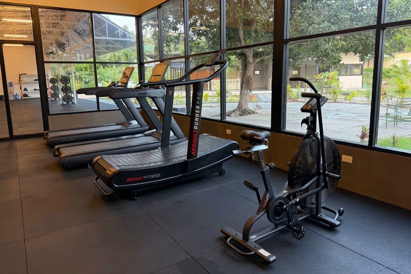 A glass-walled cardio room with curved treadmills and an air bike, floor-to-ceiling windows opening onto trees and garden outside.