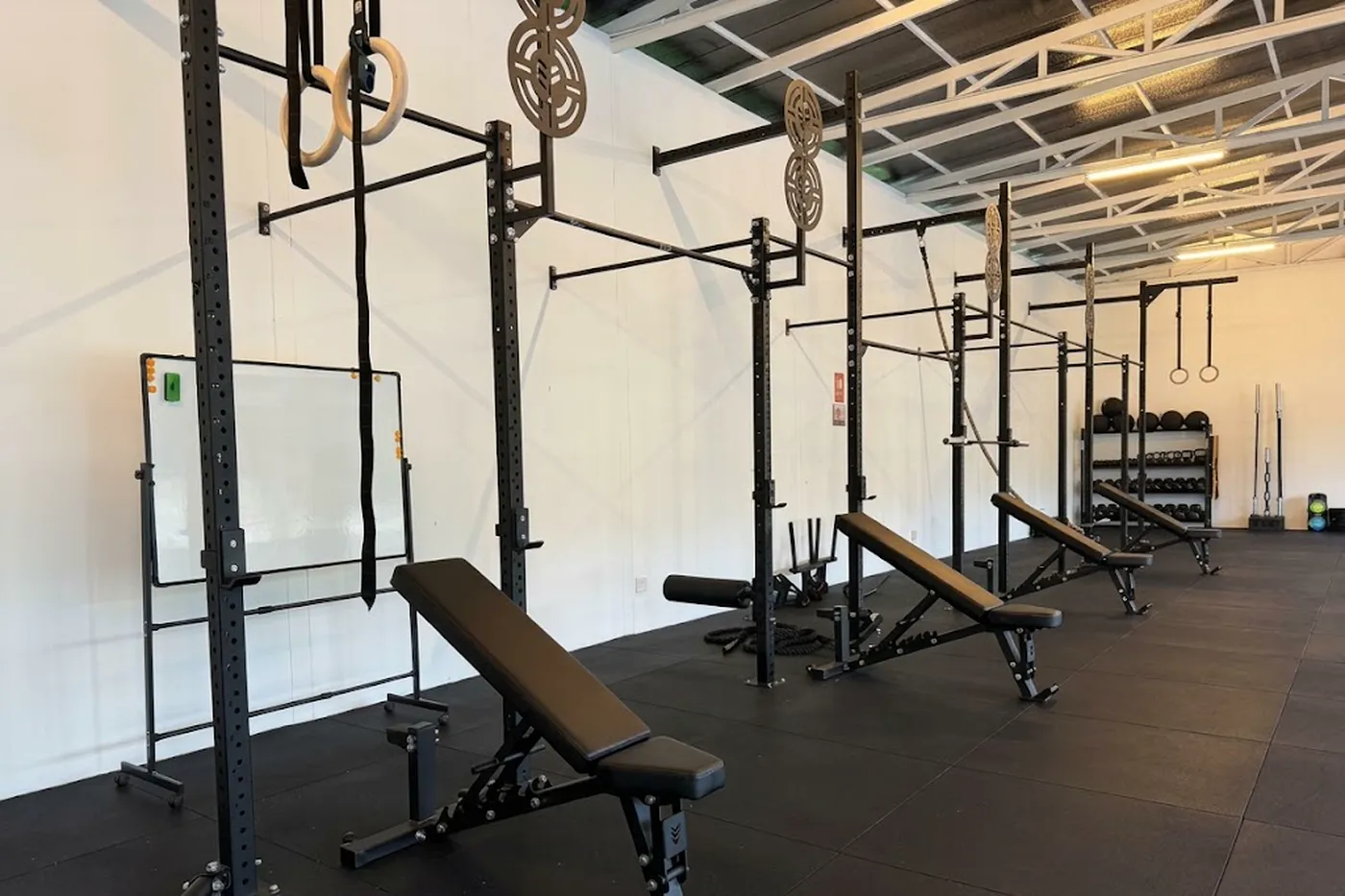 An open-air gym under a high metal roof, with squat racks, gymnastic rings and adjustable benches lined up along a white wall.