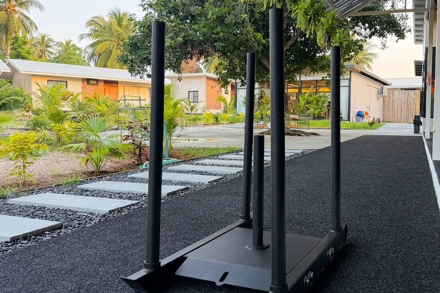 A weight sled on a rubber track lane under a covered walkway, with a tree overhead and bungalows across the garden beyond.