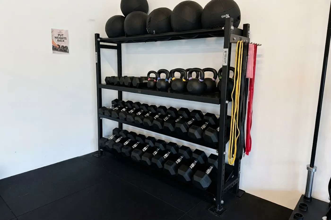 A tiered rack of dumbbells, kettlebells and medicine balls against a white wall, with resistance bands hanging at the side.