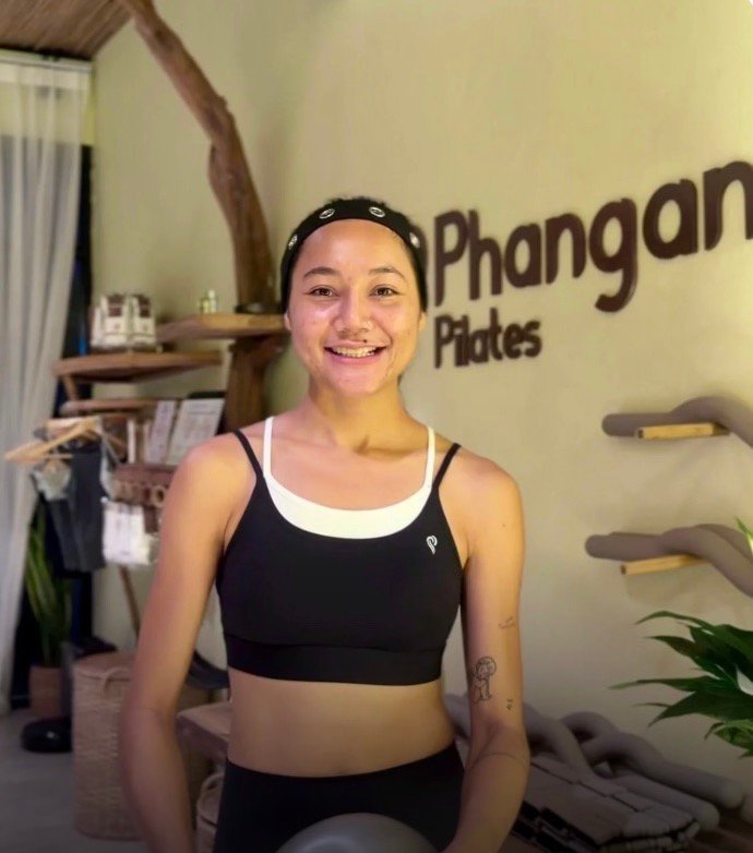 Nex, instructor at Phangan Pilates, holding a weighted Pilates ball in front of the studio's wall logo.
