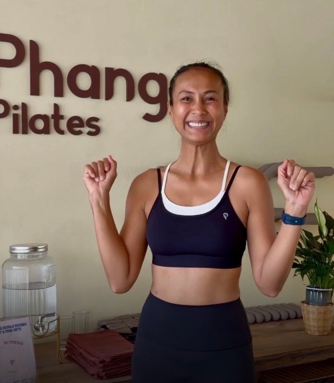 Yui, instructor at Phangan Pilates, laughing with both fists raised in front of the studio's wall logo.