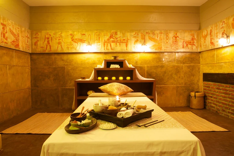 A massage table laid with a tray of oils, scrubs and rolled towels by candlelight, in a warmly lit treatment room.