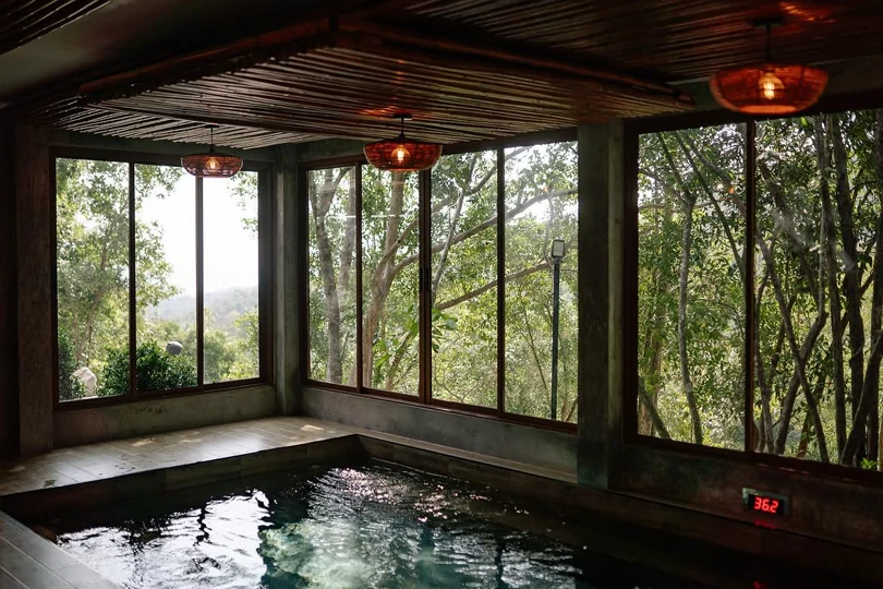 The onsen at Navana: a stone bathing pool under a slatted timber ceiling with three woven pendant lamps, glazed on two sides onto the forest.