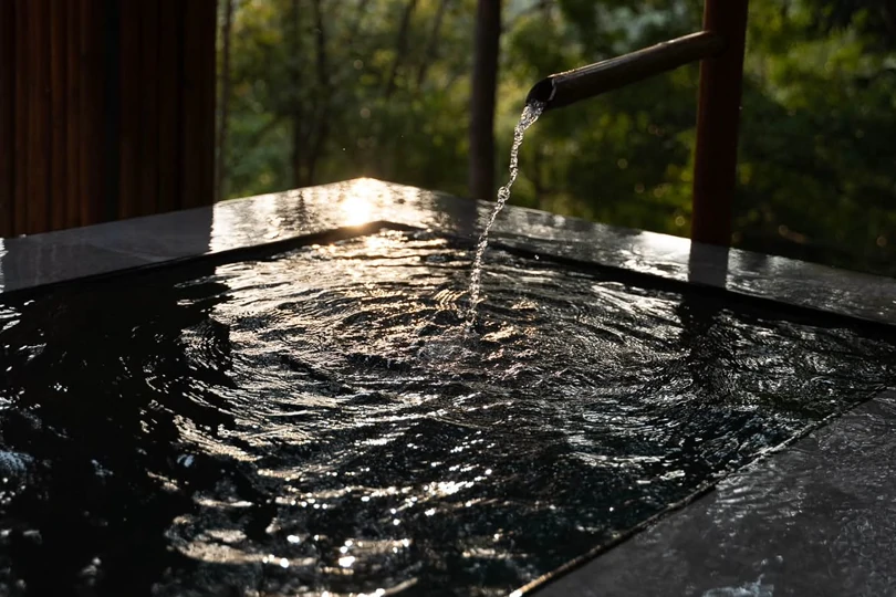 A dark stone plunge pool outdoors, water falling into it from a bamboo spout, the surface catching the light.