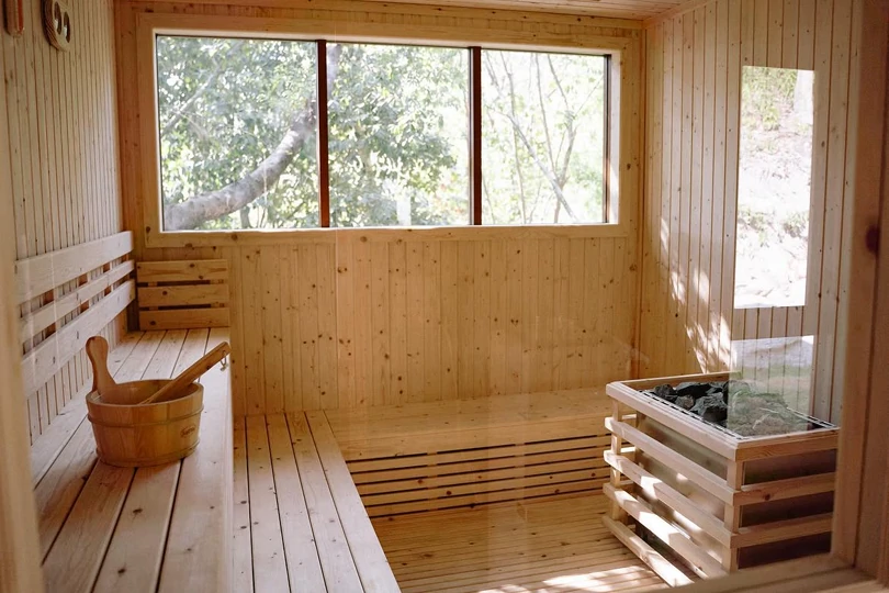 A pine sauna with tiered benches, a wooden bucket and ladle, and a stove of stones beside a window onto the trees.