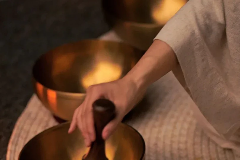 A hand striking a brass singing bowl with a wooden mallet, three more bowls arranged on a woven mat by candlelight.