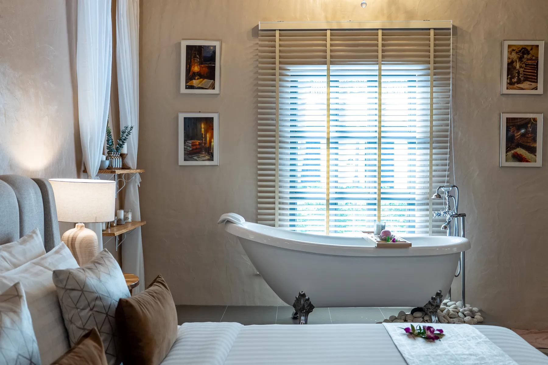 A freestanding clawfoot bathtub below a shuttered window, framed pictures on the plastered wall to either side, with the foot of the bed and folded towels in the foreground.