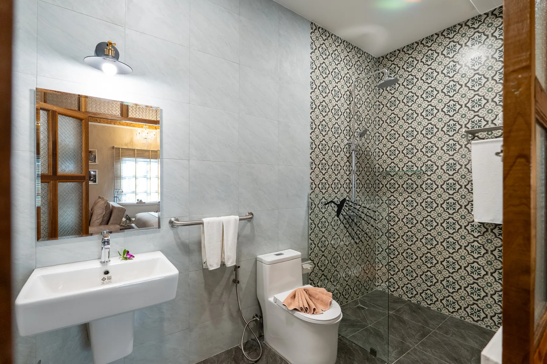 The suite bathroom: a wall-hung basin under a timber-framed mirror, folded white towels on a rail, and a walk-in rain shower against patterned encaustic tiles.