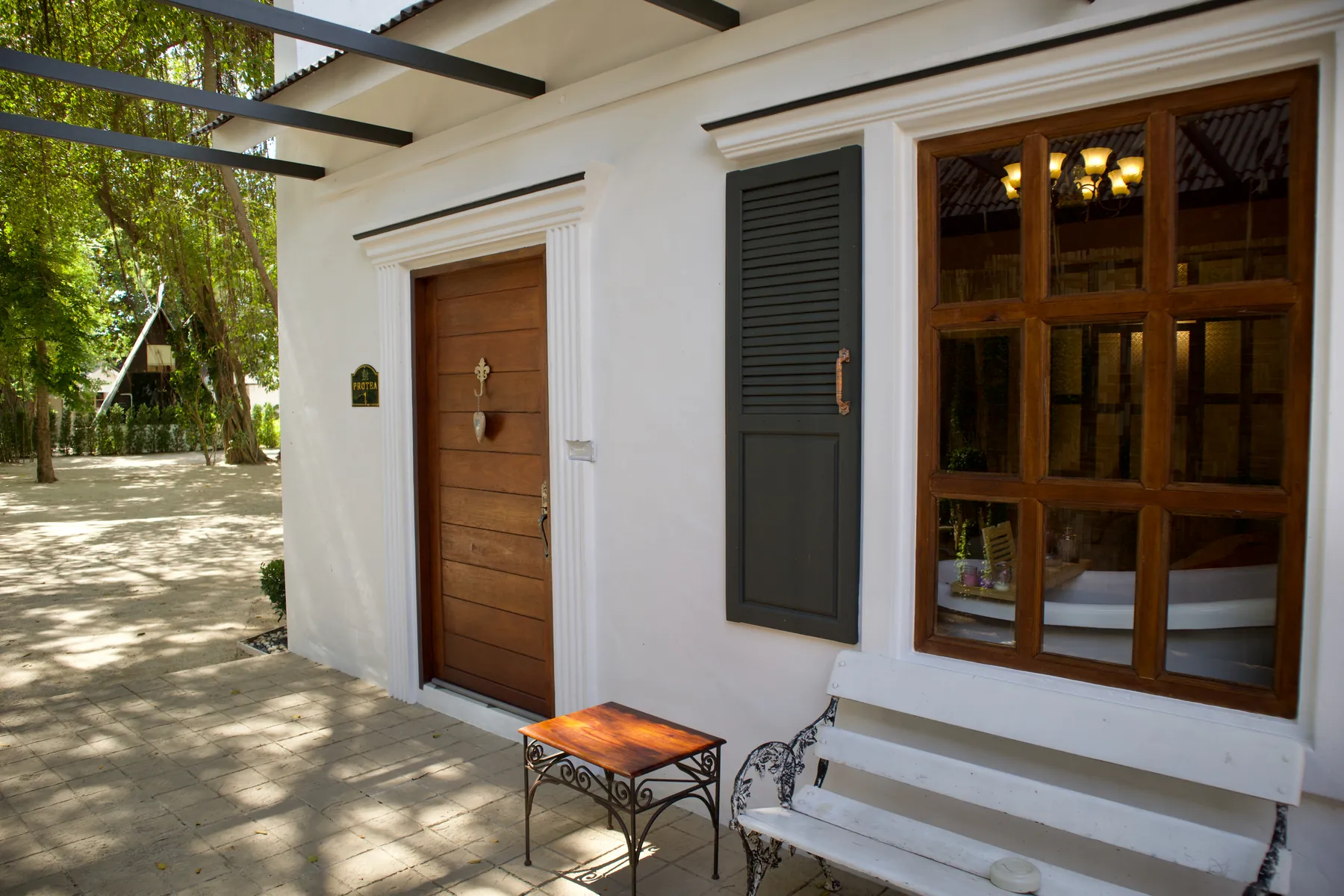 The outside of a suite at Milky Bay: a timber door and a louvred shutter in a white rendered wall under a pergola, a wrought-iron side table and a bench on the terrace, trees and dappled light beyond.