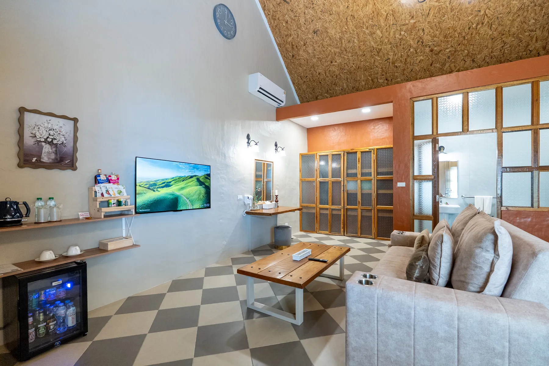 The living end of the suite: a wall-mounted television, a tea and coffee tray on open shelves, a glass-fronted minibar, a timber coffee table and a deep cushioned sofa, with the bathroom behind glazed timber doors.