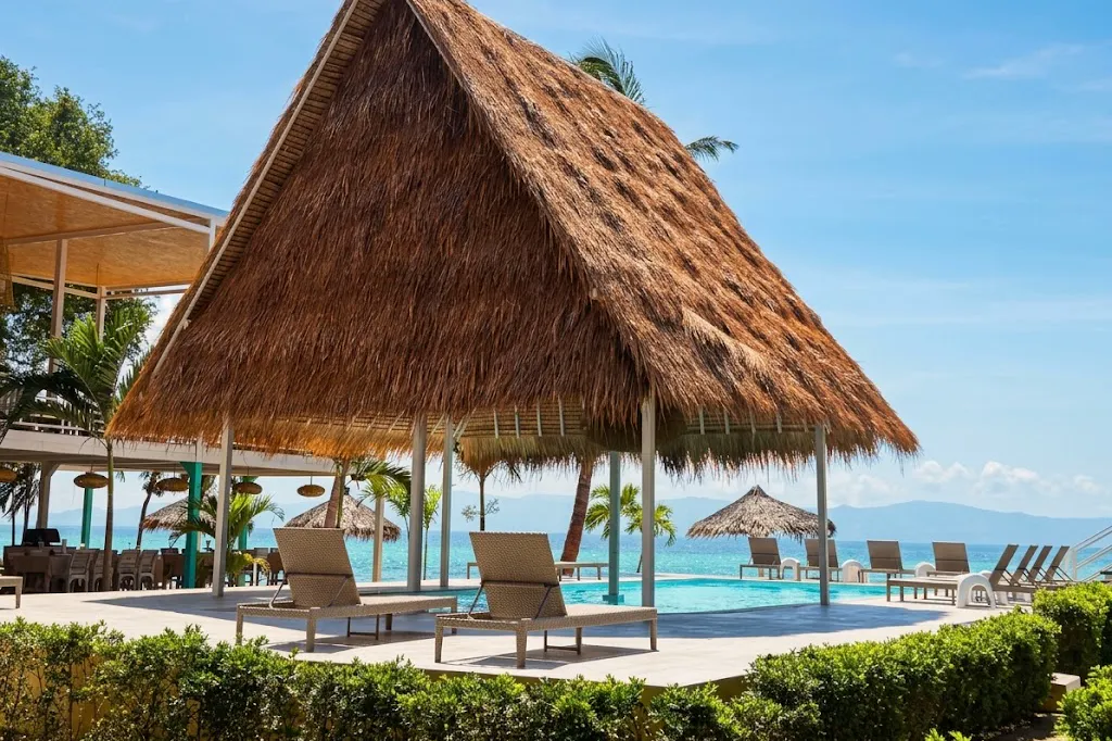 A large thatched-roof pavilion beside the pool at Milky Bay, sun loungers underneath it and the sea open behind, mountains on the horizon.