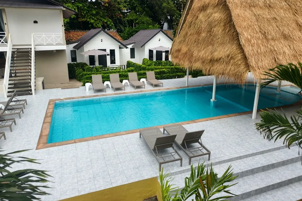 The pool at Milky Bay seen from above: a rectangular pool with sun loungers along two sides, a thatched roof to the right, and white suites with parasols behind it.