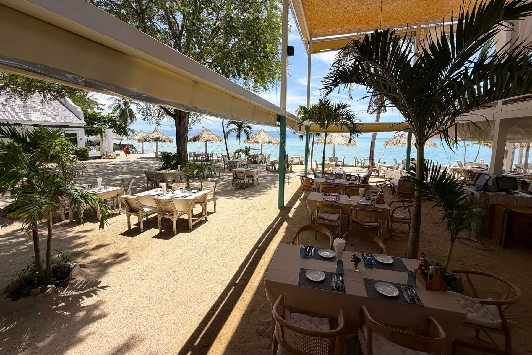 The restaurant's beachfront terrace at Milky Bay: tables laid on raked sand under a long awning, young palms planted between them, and thatched parasols at the water's edge.