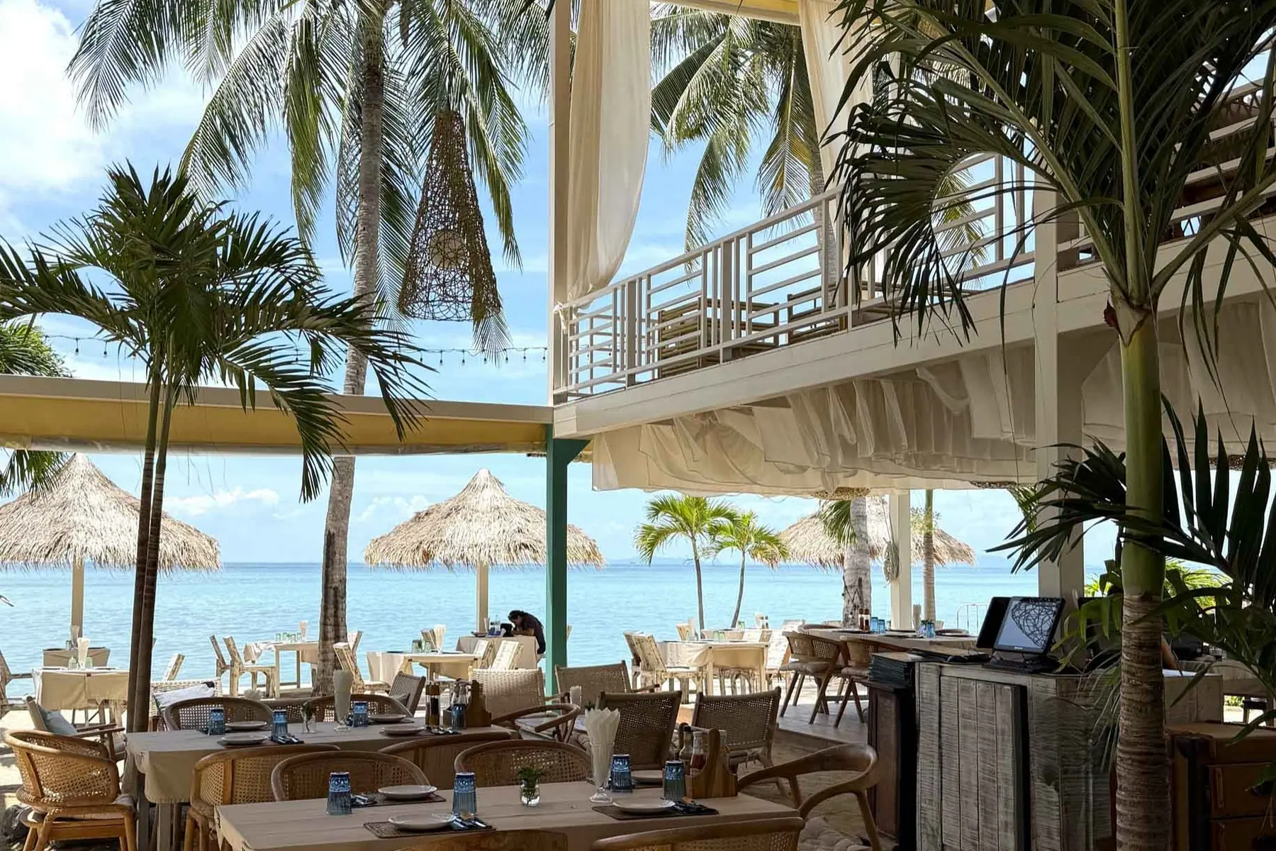 The open-sided restaurant at Milky Bay: timber tables laid for a meal, palms growing up through the room past a first-floor deck, and thatched parasols on the sand beyond with the sea behind them.