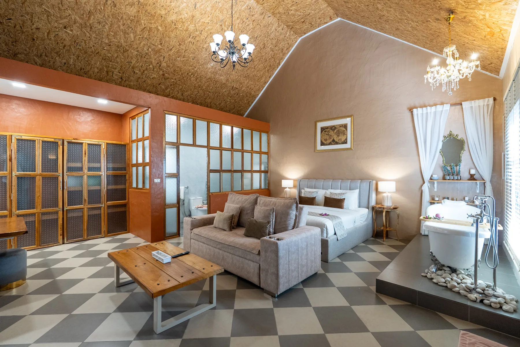A honeymoon suite at Milky Bay seen from the doorway: a sofa and low timber table on a chequerboard floor, the bed against a plastered wall, and a freestanding bathtub on a raised step by the window, under a vaulted timber ceiling with two chandeliers.