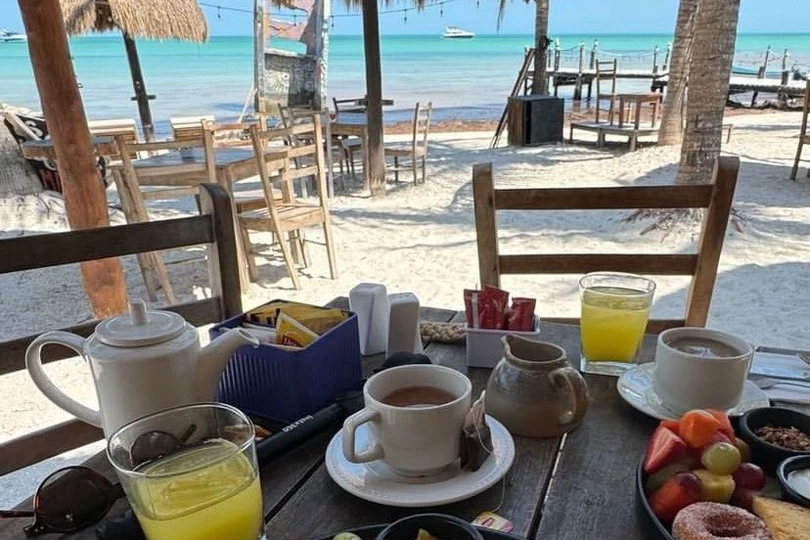 Coffee, juice and a breakfast plate on a wooden table under a thatched roof, with beach chairs, white sand and the sea beyond.