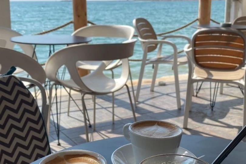 Two cups of coffee and an iced coffee on a café table beside the sea, white chairs set out on a waterfront terrace.