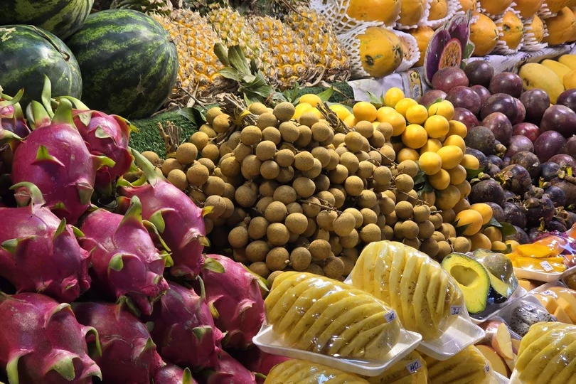 A market stall piled with whole tropical fruit: dragonfruit, longan, mango, pineapple and watermelon.