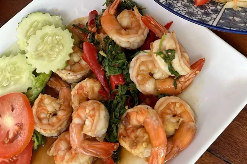 A plate of prawns stir-fried with chilli and basil, with carved cucumber and tomato beside it on a wooden table.