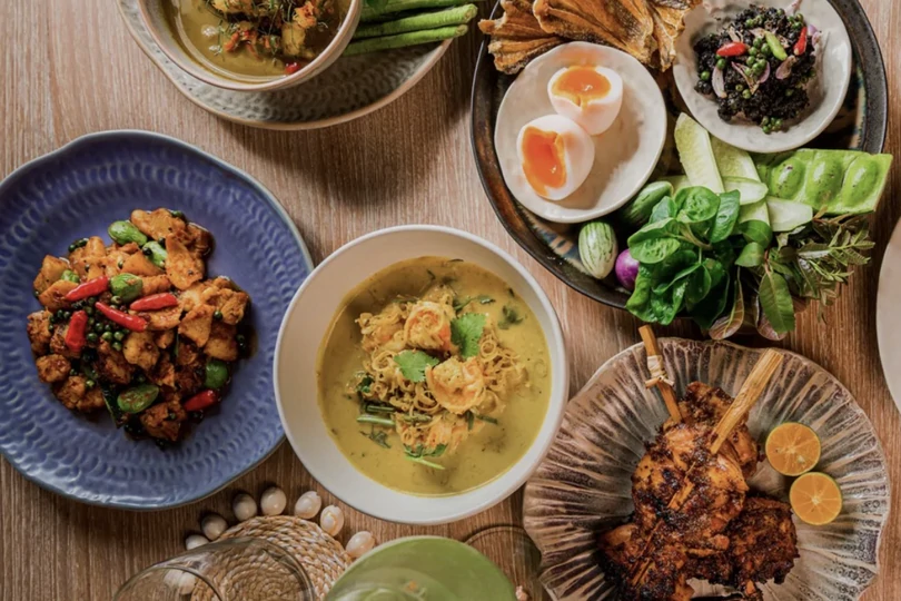 Southern Thai dishes seen from above: a curry, grilled meat, a boiled egg, fresh herbs and a cold drink on a wooden table.